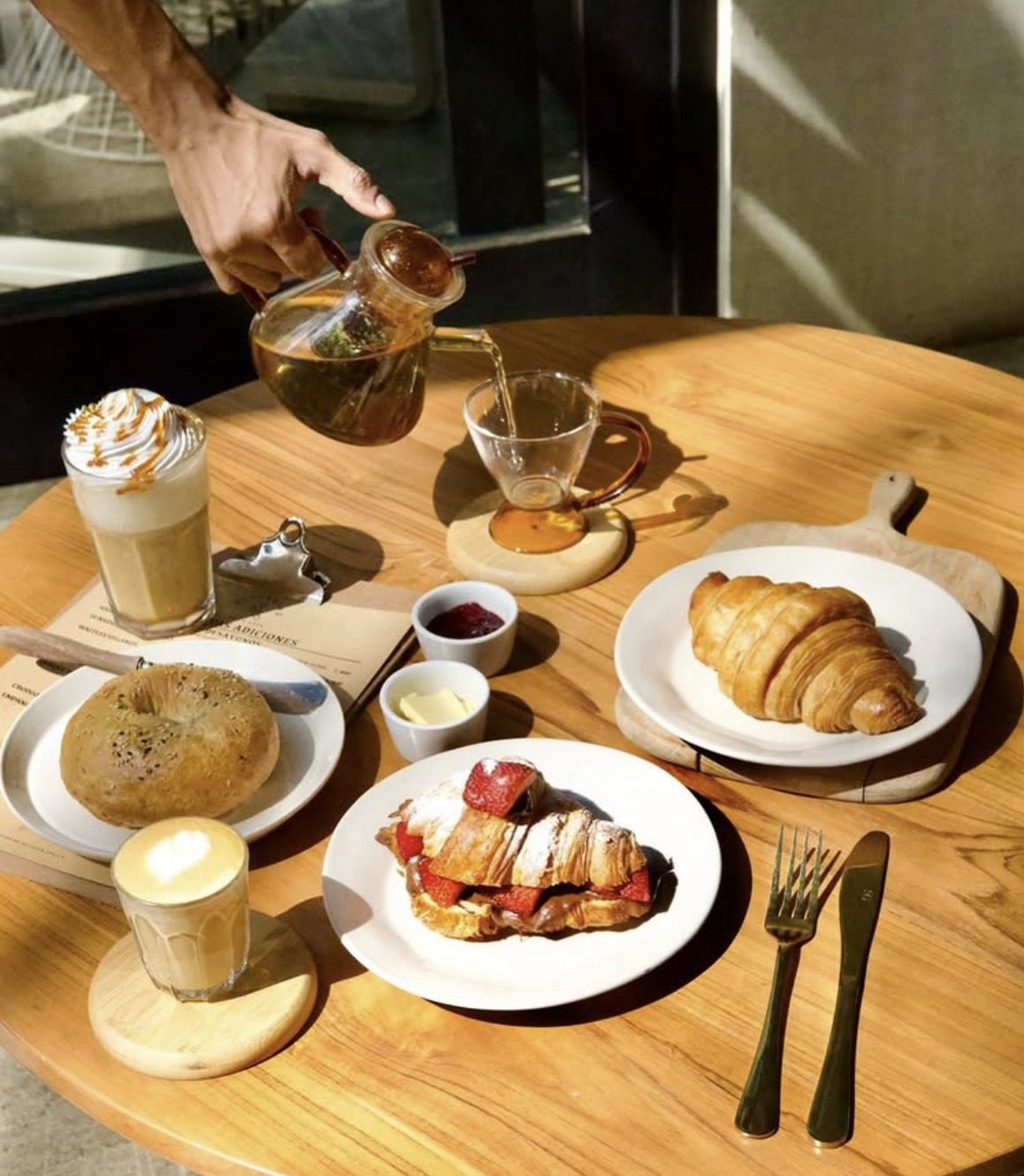 Mesa de brunch al sol con té de hierbas recién servido, capuchino con arte latte, croissant y French toast con fresas