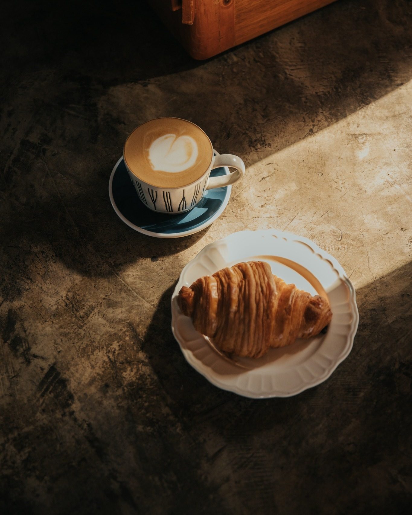 Capuchino con arte latte y croissant sobre una mesa de concreto, con un rayo de sol