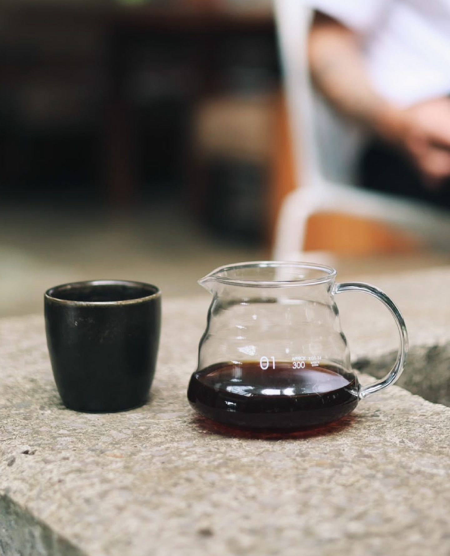 Servidor de café de método y taza oscura en la barra de concreto, con el barista al fondo
